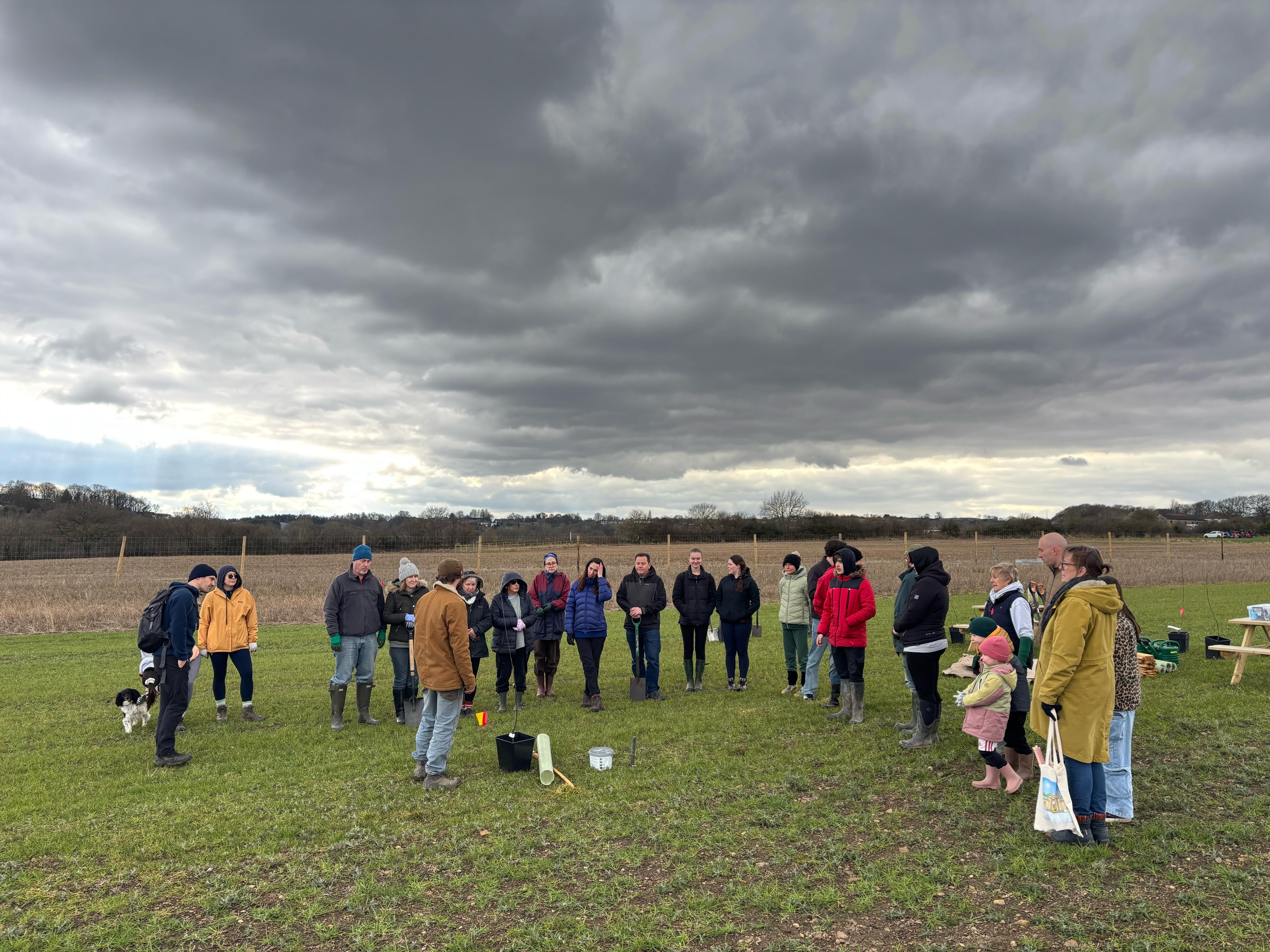 Members of the community gathering in a field on an overcast day to listen to our Patch Manager, Will, share how to plant a fruit tree in the ground